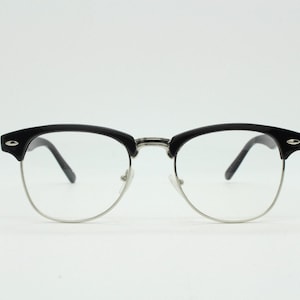Puede incluir: Gafas con montura negra en la parte superior y plateada en la inferior. Las lentes son transparentes y las patillas negras. Diseño clásico, semi-sin montura, para uso diario.