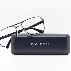 Puede incluir: Gafas rectangulares negras con puente de nariz plateado y estuche negro con la palabra "DAVIDOFF" impresa en blanco.