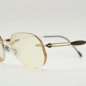 May include: Rimless eyeglasses with a gold-tone metal frame and tinted lenses. The temples are thin, with dark brown accents near the ear. The lenses have a unique, angular shape. The glasses are set against a plain white background.