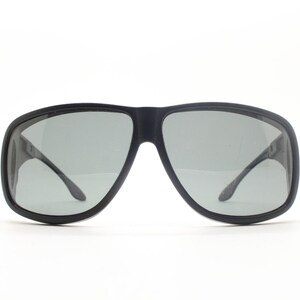 May include: Black sunglasses with gray lenses. The sunglasses have a thick, rectangular frame and curved temples. The lenses are a light gray color, and the frame is a matte black finish. The sunglasses are set against a white background.