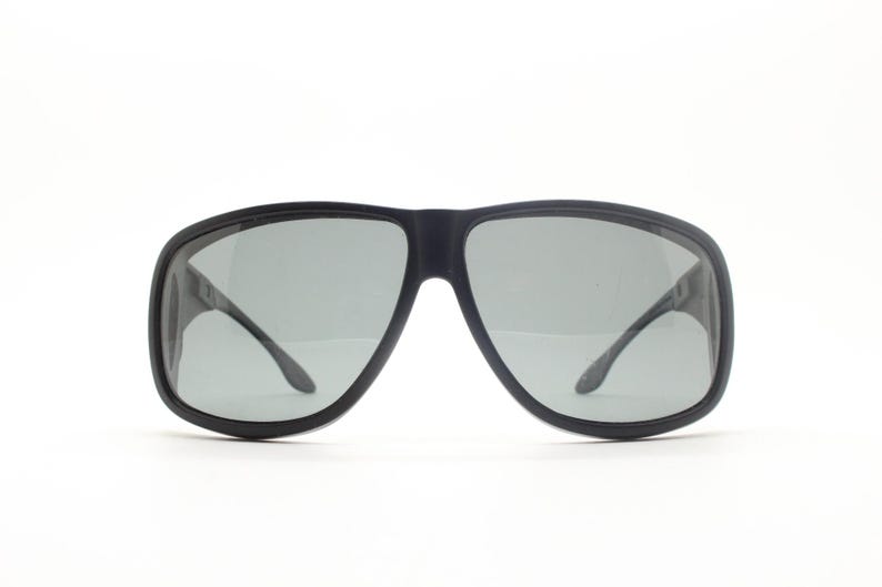 May include: Black sunglasses with gray lenses. The sunglasses have a thick, rectangular frame and curved temples. The lenses are a light gray color, and the frame is a matte black finish. The sunglasses are set against a white background.