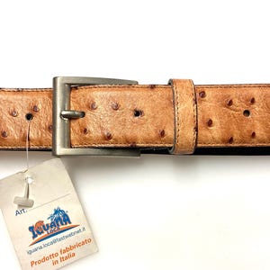 Peut inclure: Une ceinture en cuir marron clair avec une boucle argentée. La ceinture présente une surface texturée avec de petits points brun foncé. Une étiquette avec le texte "Iguana Loca" et "Prodotto fabbricato in Italia" est attachée.
