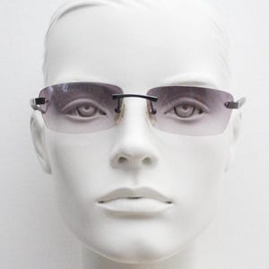 May include: Rimless sunglasses with a gradient lens. The lenses transition from a darker shade at the top to a lighter shade at the bottom. The frame is a thin, dark metal. The sunglasses are displayed on a white mannequin head.