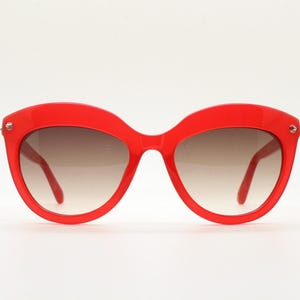 Puede incluir: Gafas de sol rojas brillantes con forma de ojo de gato y lentes degradadas marrones. Las monturas son de un rojo liso y brillante, y las patillas son finas. Las gafas tienen pequeños detalles plateados en las esquinas exteriores. Un accesorio elegante.