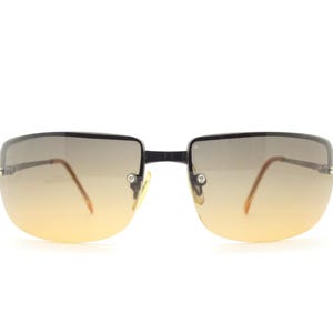 May include: Rimless sunglasses with a gradient lens that transitions from a dark gray at the top to a yellow-orange at the bottom. The frames are black and the arms are a light brown color.