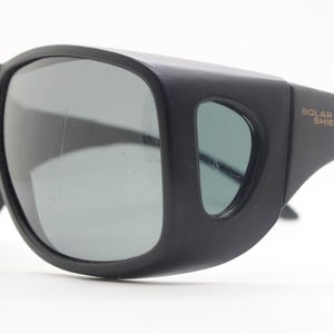 May include: Black sunglasses with wraparound frames and gray lenses. The sunglasses have a thick, sturdy design with the words "SOLAR SHIELD" printed on the arm.