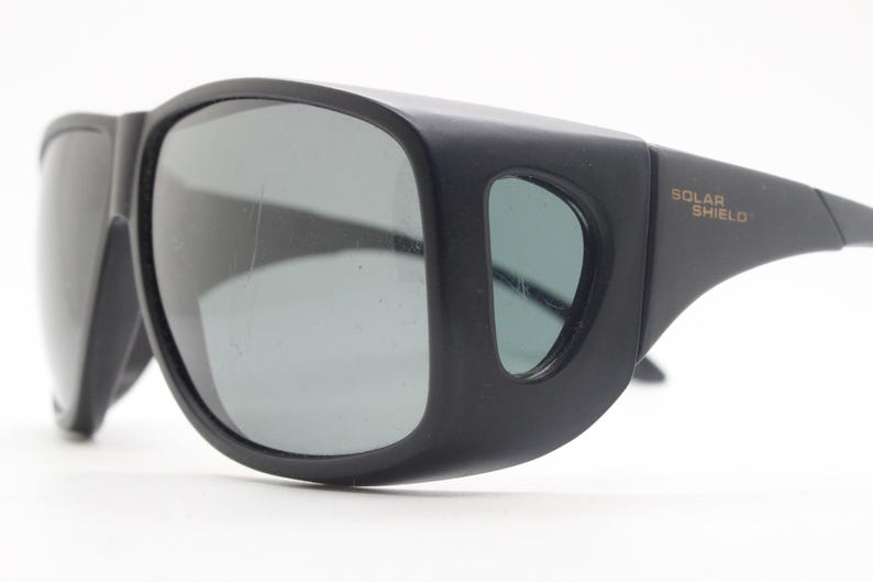 May include: Black sunglasses with wraparound frames and gray lenses. The sunglasses have a thick, sturdy design with the words "SOLAR SHIELD" printed on the arm.