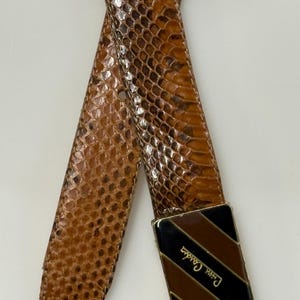 Puede incluir: Un cinturón de piel de serpiente marrón con una hebilla dorada. La hebilla tiene un diseño a rayas negras y marrones con el logotipo de Pierre Cardin. El cinturón está hecho de piel de serpiente.