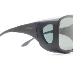 May include: Black sunglasses with large, square lenses and side shields. The text "SOLAR SHIELD" is printed on the temple. The sunglasses have a curved arm and a light green tint to the lenses.
