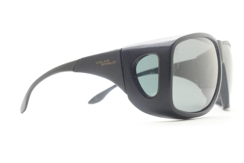 May include: Black sunglasses with large, square lenses and side shields. The text "SOLAR SHIELD" is printed on the temple. The sunglasses have a curved arm and a light green tint to the lenses.