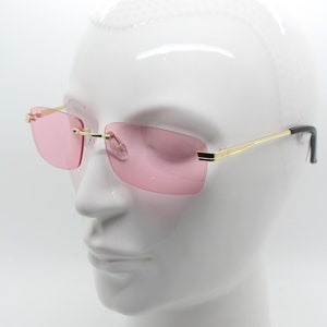 May include: Rimless sunglasses with pink lenses and gold-colored frames. The temples are gold with black tips. The sunglasses are displayed on a white mannequin head.