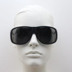 May include: Black sunglasses with a thick, rectangular frame. The sunglasses have dark lenses and are displayed on a white mannequin head. The overall style is modern and bold.
