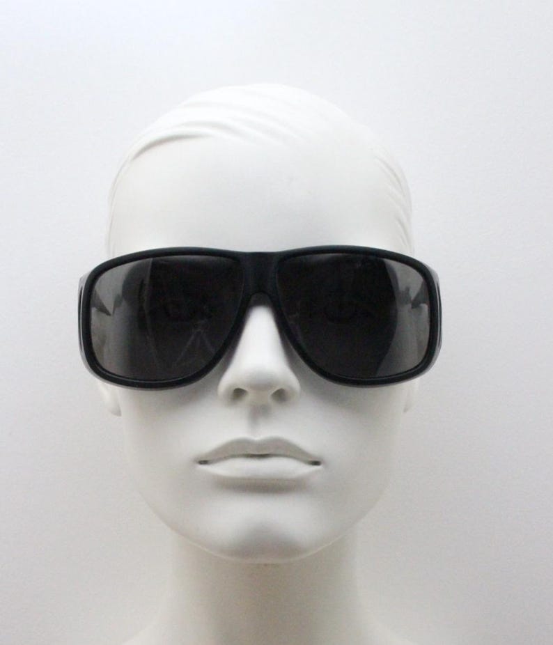 May include: Black sunglasses with a thick, rectangular frame. The sunglasses have dark lenses and are displayed on a white mannequin head. The overall style is modern and bold.