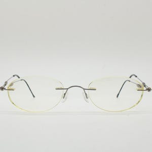 May include: Rimless eyeglasses with oval lenses and thin, silver-toned metal frames. The temples are black and curve around the ears. The glasses have a minimalist design, suitable for everyday wear.