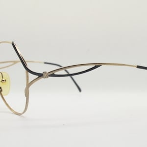 Könnte beinhalten: Goldfarbene Metallbrille mit schwarzen Akzenten an den Bügeln. Die Brille hat ein einzigartiges, geschwungenes Design.
