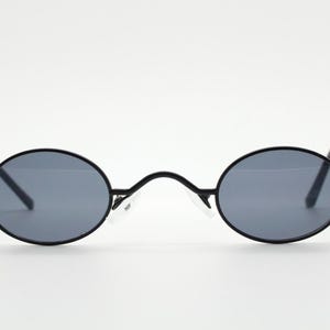 May include: Black oval sunglasses with gray lenses. The sunglasses have a thin black frame and temples. The bridge of the sunglasses has a curved design. The sunglasses are set against a white background.