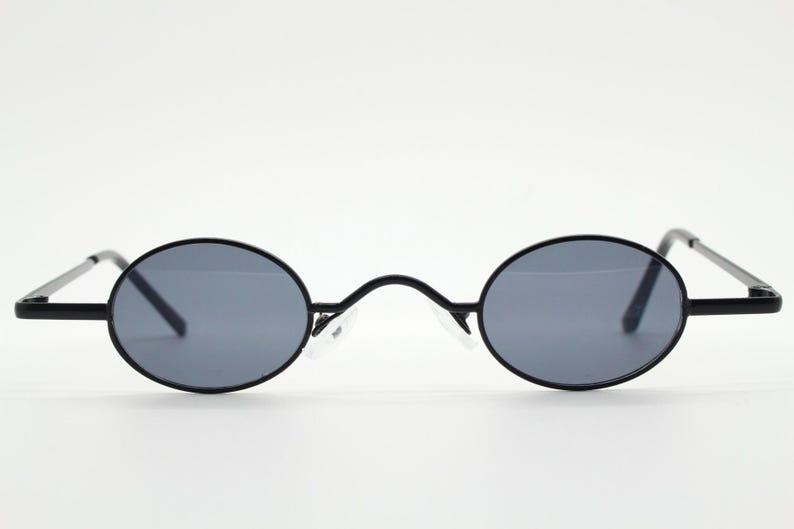 May include: Black oval sunglasses with gray lenses. The sunglasses have a thin black frame and temples. The bridge of the sunglasses has a curved design. The sunglasses are set against a white background.