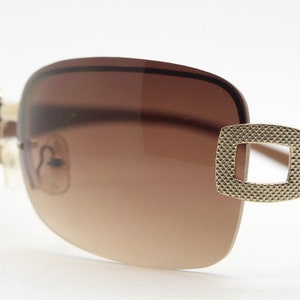 May include: Brown sunglasses with a gold-tone frame and a textured detail on the side. The lenses are brown and gradient.