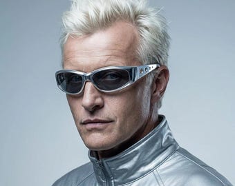 May include: A pair of silver-toned wraparound sunglasses with dark lenses and perforated side details, worn by an individual with short, spiky platinum blonde hair and a high-collared metallic silver jacket against a neutral gray background.