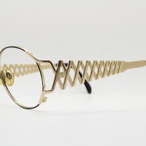 May include: Gold-toned eyeglasses with a unique geometric design. The frames feature a series of diamond-shaped cutouts along the temples, with black earpieces. The lenses are oval-shaped, and the bridge has a delicate, open design.