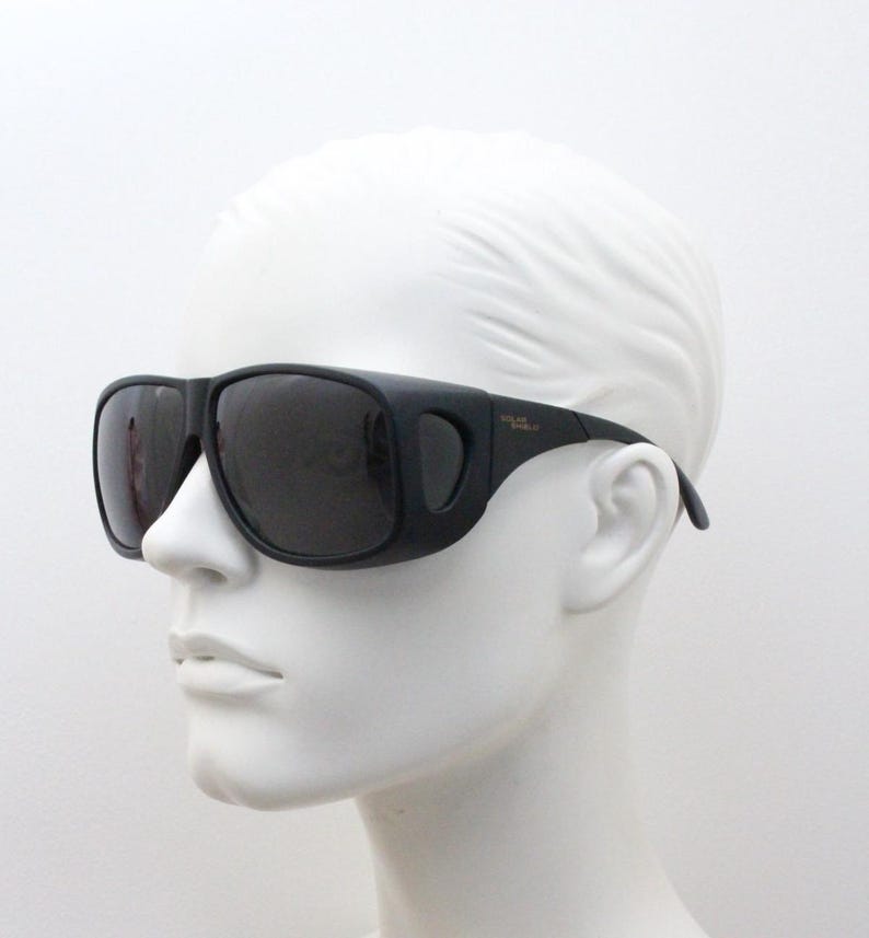 May include: Black sunglasses with a wraparound design, featuring dark lenses and a thick frame. The sunglasses are displayed on a white mannequin head, showcasing their shape and style. The text "POLAR SHIELD" is visible on the temple.