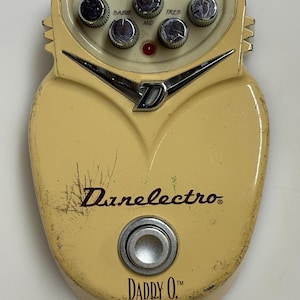 Puede incluir: Un pedal de efectos para guitarra Danelectro Daddy O. de color crema con forma redondeada de escudo. Cuenta con cinco perillas de control cromadas etiquetadas como VOL, BASS, MID, TREB y O. DRIVE, un interruptor de pie plateado y un logo D cromado.