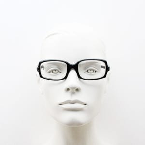 May include: A pair of black rectangular eyeglasses with clear lenses positioned on a white mannequin head. The frames feature a solid black finish with a bridge connecting the two lenses and thin temples extending along the sides of the head.