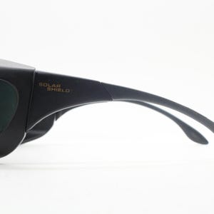 May include: Black Solar Shield sunglasses with a wraparound design. The glasses have dark green lenses and curved temples for a secure fit. The text "SOLAR SHIELD" is printed on the temple.