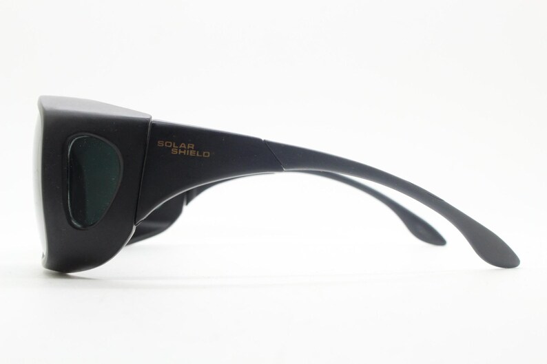 May include: Black Solar Shield sunglasses with a wraparound design. The glasses have dark green lenses and curved temples for a secure fit. The text "SOLAR SHIELD" is printed on the temple.