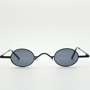 May include: Oval-shaped sunglasses with a black frame and gray lenses. The sunglasses have a thin frame and straight temples. The lenses are a dark gray color, and the bridge is a small, clear plastic piece.