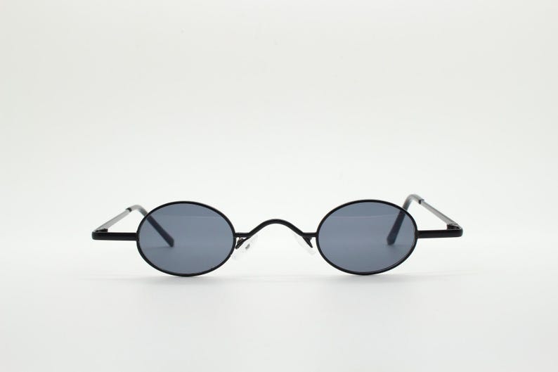 May include: Oval-shaped sunglasses with a black frame and gray lenses. The sunglasses have a thin frame and straight temples. The lenses are a dark gray color, and the bridge is a small, clear plastic piece.