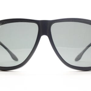 May include: Black sunglasses with gray lenses. The sunglasses have a thick, rounded frame and a bridge connecting the two lenses. The temples are thin and curve around the ears. The lenses are a light gray color.