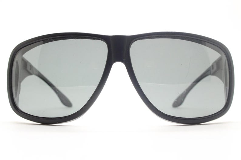 May include: Black sunglasses with gray lenses. The sunglasses have a thick, rounded frame and a bridge connecting the two lenses. The temples are thin and curve around the ears. The lenses are a light gray color.