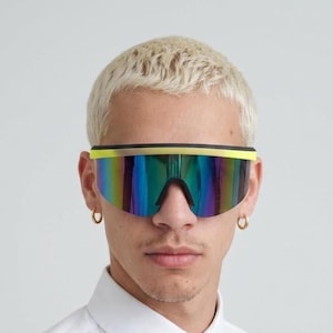 Puede incluir: Gafas de sol tipo máscara de gran tamaño con una lente única continua de acabado iridiscente reflectante que cambia entre tonos azules, verdes y morados. La montura presenta una barra superior en amarillo neón y estructura negra.