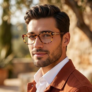 May include: Tortoiseshell eyeglasses with a rectangular frame shape and clear lenses. The frames feature a distinct brown and amber mottled pattern with metallic accents on the temples, worn over a white collared shirt and a rust-colored jacket.