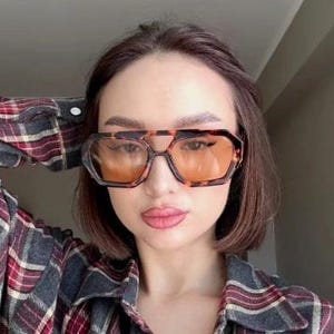 Puede incluir: Gafas de sol con montura de carey y lentes naranjas. Las gafas tienen un doble puente y una forma geométrica. La persona lleva una camisa a cuadros en rojo, negro y blanco.