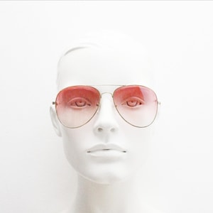 May include: Aviator sunglasses featuring thin gold-tone metal frames and gradient lenses that transition from a deep pink at the top to a lighter, translucent pink at the bottom, worn on a white mannequin head against a plain white background.