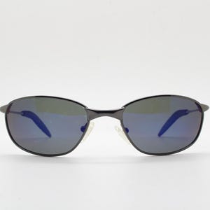 May include: Black oval sunglasses with blue mirrored lenses. The sunglasses have a dark frame and a white nose bridge. The lenses reflect a blue hue. The sunglasses are set against a white background.