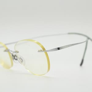 May include: Rimless eyeglasses with oval lenses and silver-toned metal frames. The lenses have a slight yellow tint. The temples are thin and curve behind the ears. The glasses are set against a white background.