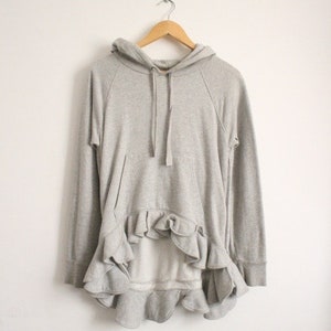 May include: A grey hooded sweatshirt with a ruffled hem. The sweatshirt has long sleeves and a drawstring hood.