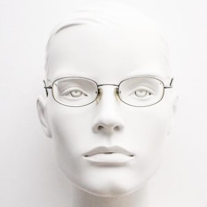 May include: Oval-shaped eyeglasses with a thin, dark metal frame. The glasses are displayed on a white mannequin head, showcasing the minimalist design. The lenses are clear, and the overall aesthetic is understated and classic.