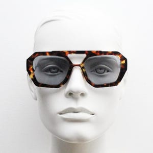 Puede incluir: Gafas de sol con estampado de carey y montura geométrica. Las gafas tienen un diseño marrón y amarillo con lentes grises. Se muestran sobre una cabeza de maniquí blanca.