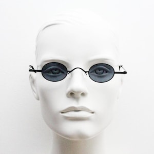 May include: Oval sunglasses with dark lenses and a slender, black metal frame. The glasses are presented on a white mannequin head, highlighting the design. The lenses have a dark grey tint. The frame is a thin wire.