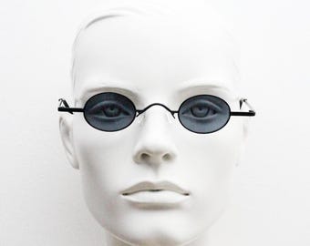 May include: Oval-shaped sunglasses with dark lenses and a thin, black metal frame. The sunglasses are displayed on a white mannequin head, showcasing the eyewear's design and shape. The lenses appear to be a dark gray color.