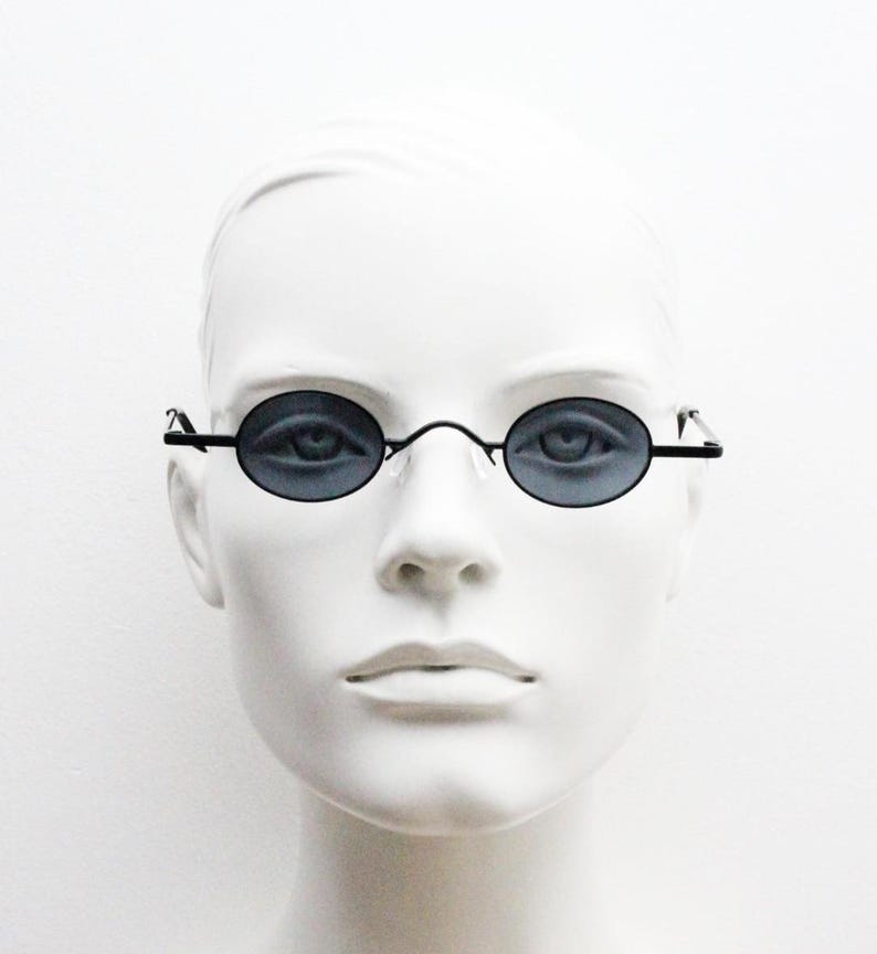 May include: Oval-shaped sunglasses with dark lenses and a thin, black metal frame. The sunglasses are displayed on a white mannequin head, showcasing the eyewear's design and shape. The lenses appear to be a dark gray color.