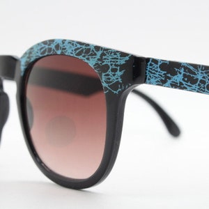 Vintage ronde zonnebril met wayfarer-design uit de jaren 80. Blauw voorhoofd en armen met abstract patroon op een zwart montuur met glazen met verloop. Ongebruikte NOS