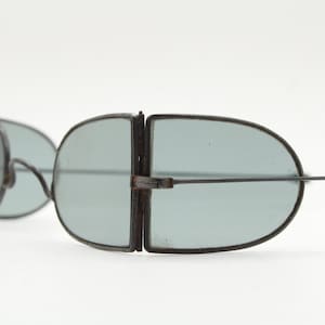 May include: Vintage sunglasses with a unique design. The lenses are a light blue-green color, set in a dark metal frame. The frames have a distinctive shape, with a hinge in the middle. The arms are thin and extend to the sides.