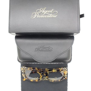 Puede incluir: Un conjunto de gafas con gafas con estampado de carey y cristales, sobre un paño negro. El conjunto incluye un estuche de cuero negro y una caja negra, ambos con el logotipo "Agent Provocateur".