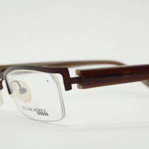 May include: Brown rectangular eyeglasses with clear lenses. The frames are a dark brown colour with a rectangular shape. The temples are a lighter brown. The brand name "WILLIAM MORRIS LONDON" is printed on the lens.