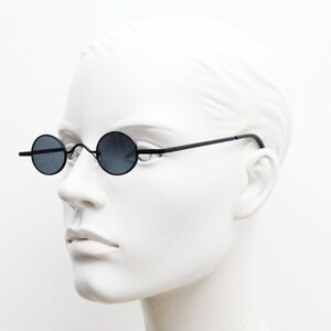 May include: Black round sunglasses with thin black frames. The lenses are dark, and the glasses have a minimalist design. The sunglasses are displayed on a white mannequin head.